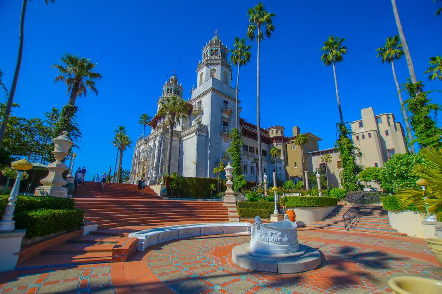 Hearst Castle rises above a grand terraced courtyard under a cloudless blue California sky. The white Mediterranean Revival-style mansion features ornate twin bell towers, decorative stonework, and red-tiled roofs. Palm trees frame the scene, while broad brick staircases, manicured gardens, and an elegant marble fountain lead visitors toward the historic estate.