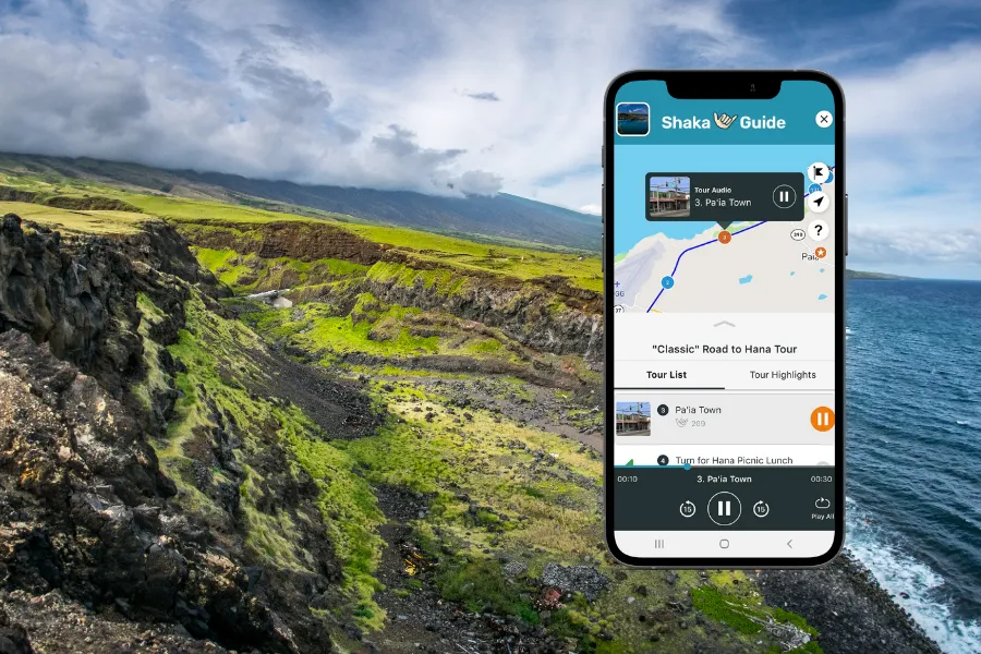 Smartphone displaying the Shaka Guide app over a dramatic coastal cliff landscape with green hills, rocky terrain, and ocean views.