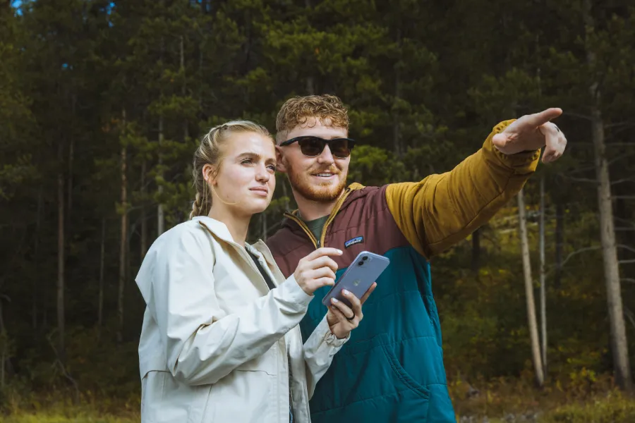 Two travelers stand in front of a forest holding a phone, while one points toward something in the distance.