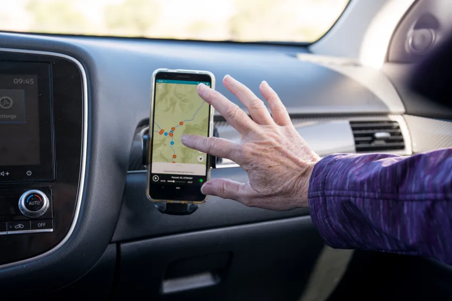 Hand using a smartphone navigation app mounted on a car dashboard, with the interior controls and windshield visible nearby.