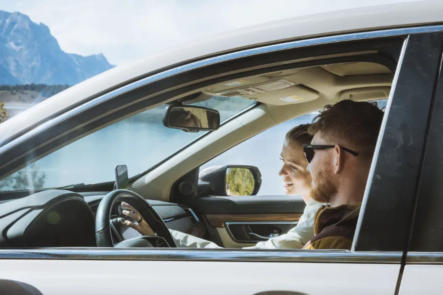 Two travelers sit inside a car beside a mountain lake, enjoying the scenic view through the open window.