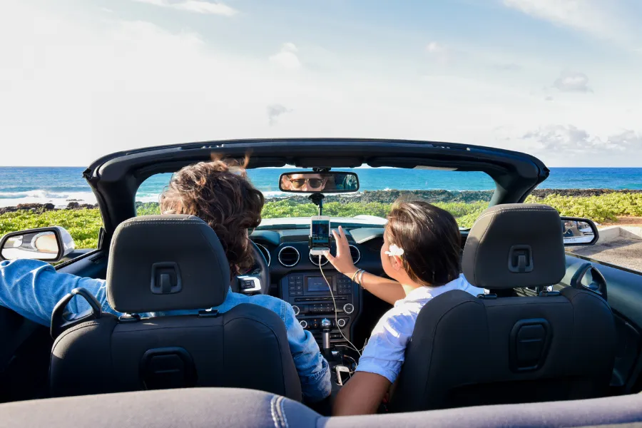 Two travelers sit in a convertible by the ocean, using a smartphone navigation app while looking out at the coastal view.