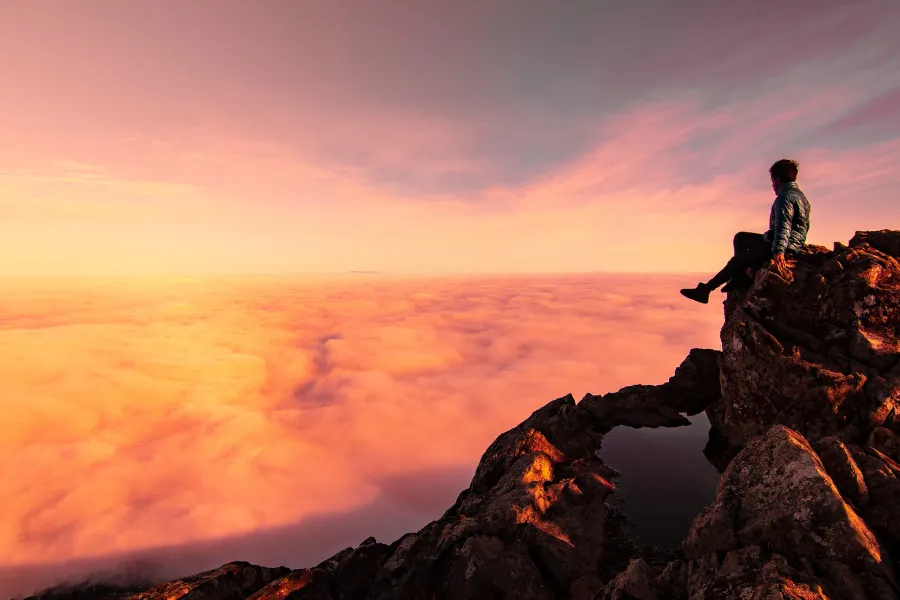 Traveler sits on a rocky mountain peak above a sea of clouds glowing pink and orange at sunrise.