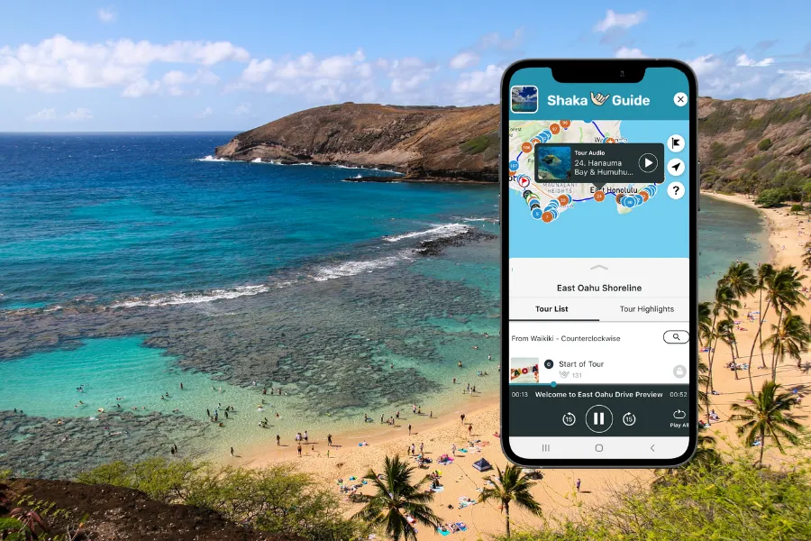 Smartphone displaying the Shaka Guide app over a tropical beach cove with turquoise water, sandy shore, and rocky coastline.