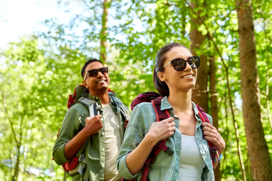 Two hikers wearing backpacks and sunglasses walk through a sunlit forest with bright green trees all around.