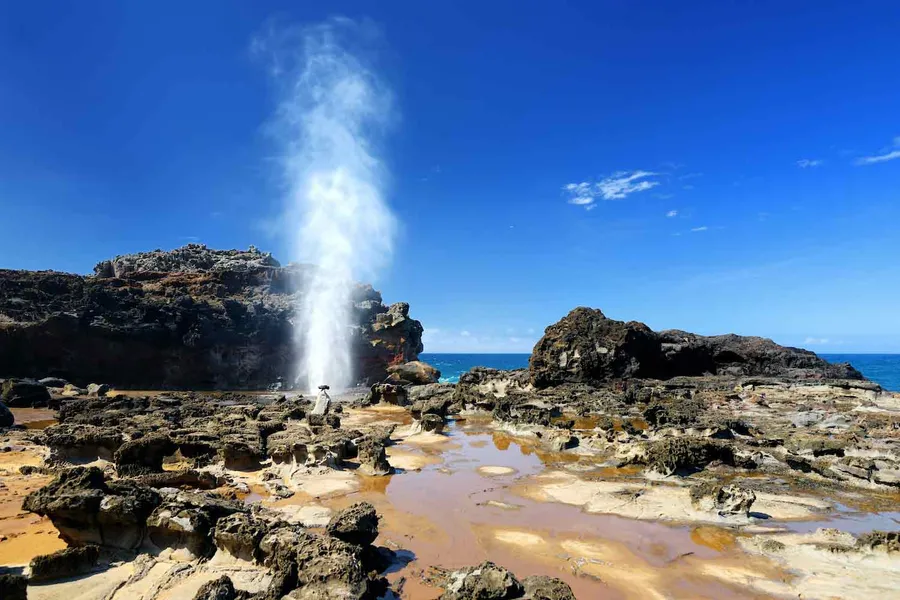 Blowhole shooting a tall plume of water above a rocky coastal shoreline under a clear blue sky.