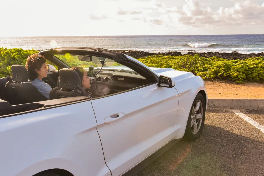 Two travelers sit in a white convertible parked by the ocean at sunset, with waves, coastal greenery, and warm golden light in the background.