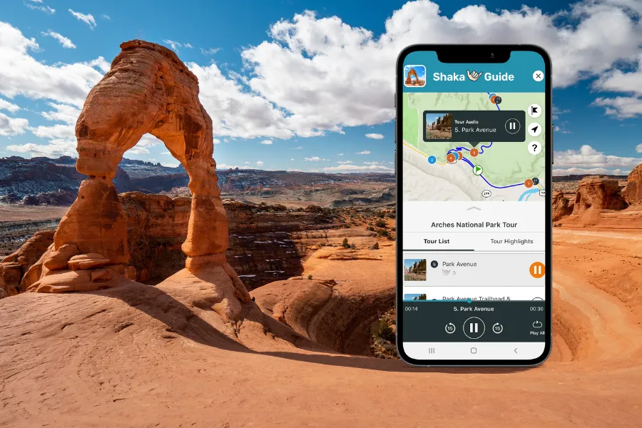 Smartphone displaying the Shaka Guide app beside Delicate Arch in a red rock desert landscape under a bright blue sky with clouds.
