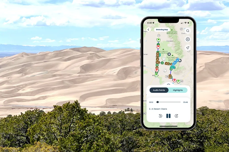 Smartphone displaying an audio tour map of Great Sand Dunes National Park, with vast sand dunes in the background under a partly cloudy sky.