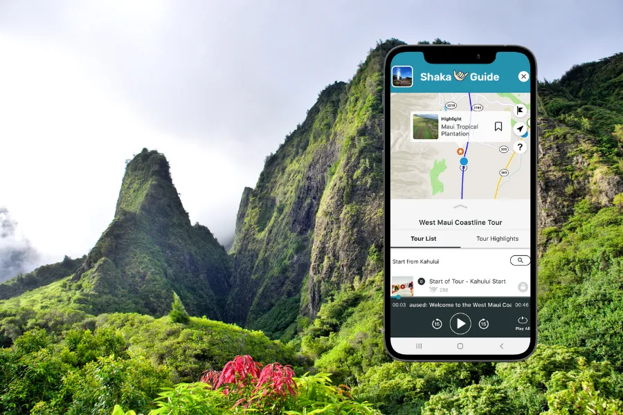 Smartphone displaying the Shaka Guide app over lush green mountain peaks and dense tropical vegetation in a dramatic valley landscape.