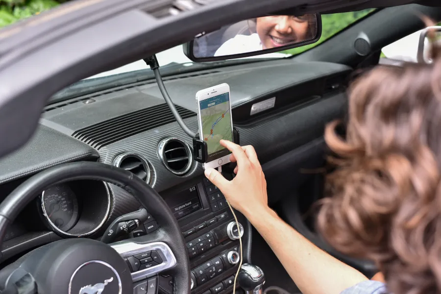 Driver using a smartphone navigation app mounted on the dashboard inside a car, with the route reflected in the rearview mirror.