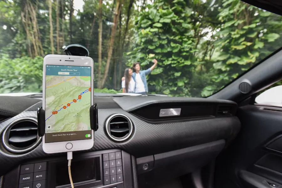 Smartphone displaying a driving tour map inside a car, with two travelers taking a selfie outside in a lush tropical forest.