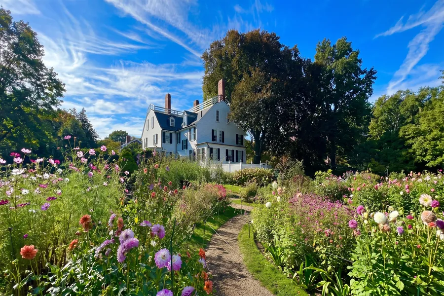 A gravel path winds through a vibrant cottage garden bursting with dahlias and cosmos in shades of pink, peach, and white, leading to a stately white Colonial-era house framed by mature trees under a brilliant blue sky.