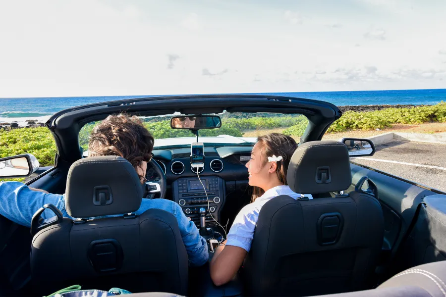 Two travelers sit in a convertible beside the ocean, with a smartphone navigation app mounted on the dashboard and a coastal view ahead.