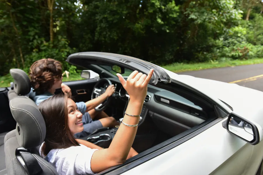 Two travelers ride in a convertible along a lush tropical road, with one passenger smiling and throwing a shaka hand sign.
