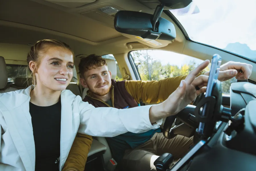 Two travelers sit inside a car while adjusting a mounted smartphone, with one pointing ahead during the drive.
