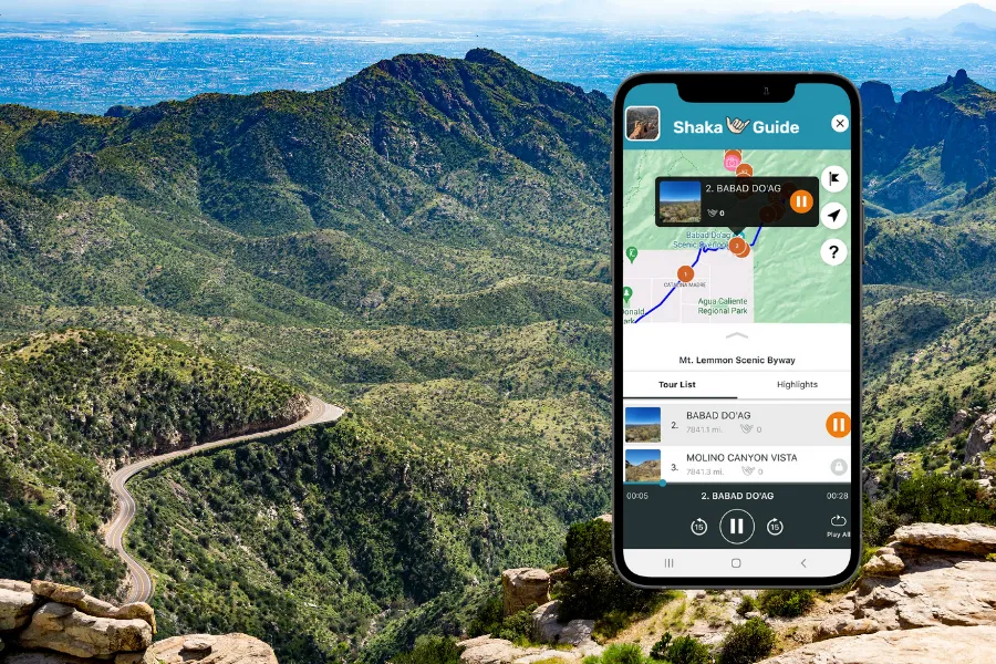 Smartphone displaying the Shaka Guide app over a sweeping mountain landscape with a winding road cutting through green desert hills below.