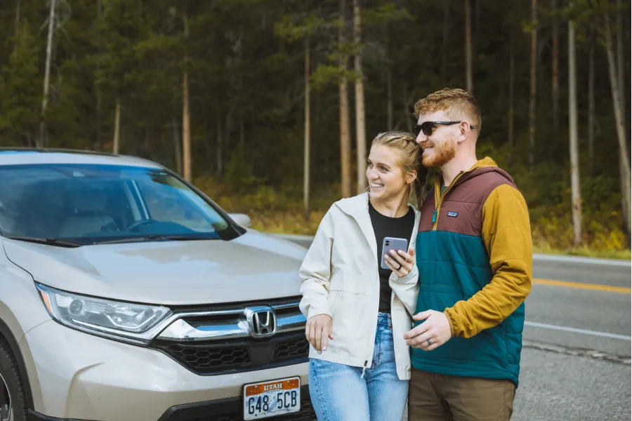 Two travelers stand smiling beside a parked SUV on a forest road, holding a phone and looking off into the distance.
