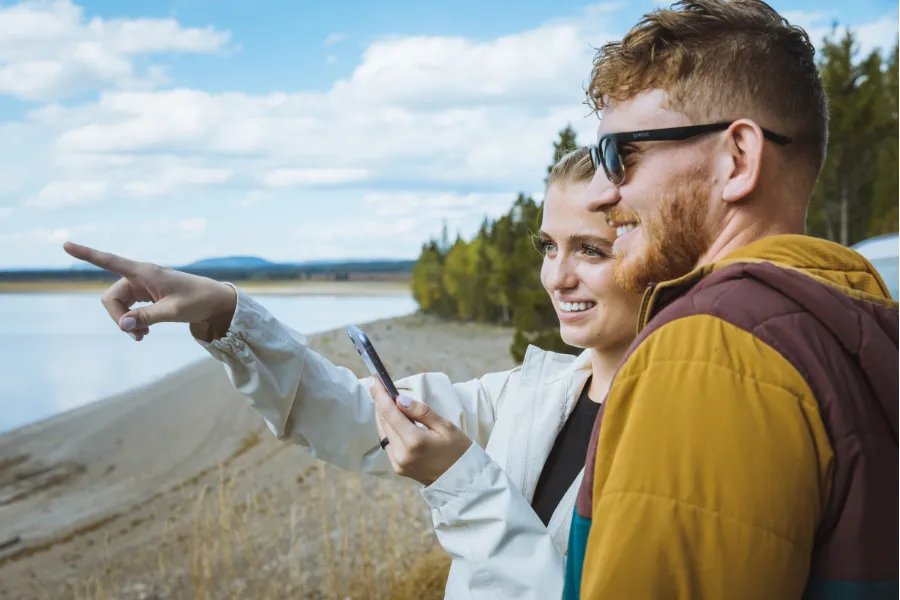 Two travelers stand by a lakeshore with a phone, smiling as one points toward the distant view.