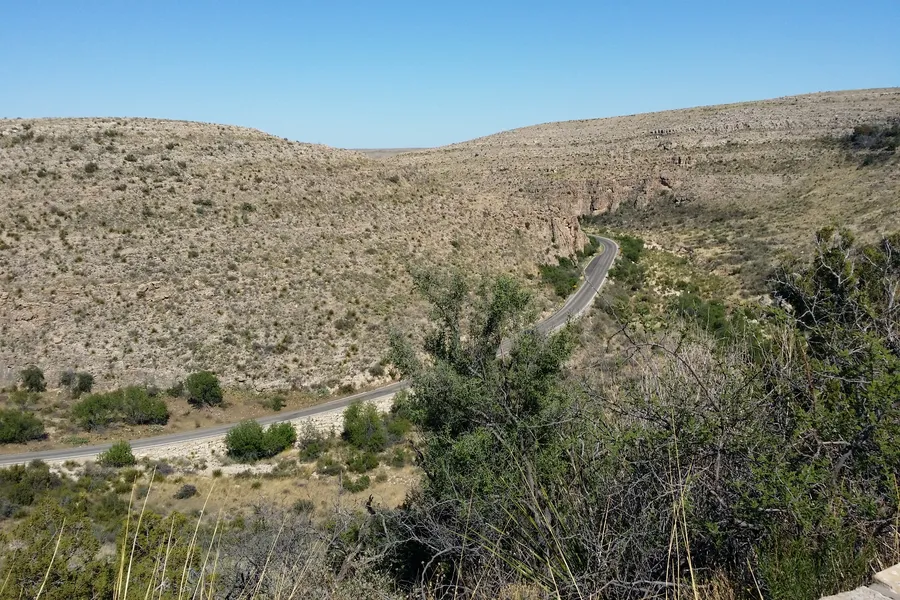 A narrow two-lane road winds through the arid, scrub-covered hills of Walnut Canyon Vista near Carlsbad Caverns National Park under a clear blue sky.