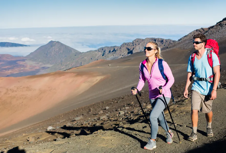 Two hikers walk across a rugged volcanic trail with trekking poles, backpacks, and expansive mountain views above the clouds.