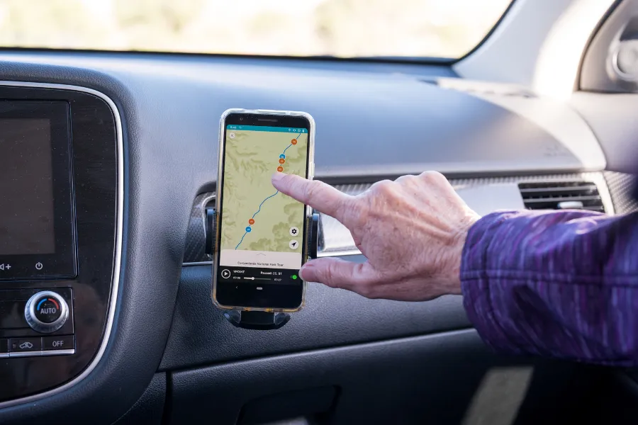 Hand pointing to a smartphone tour map mounted on a car dashboard, with the vehicle’s interior visible around it.
