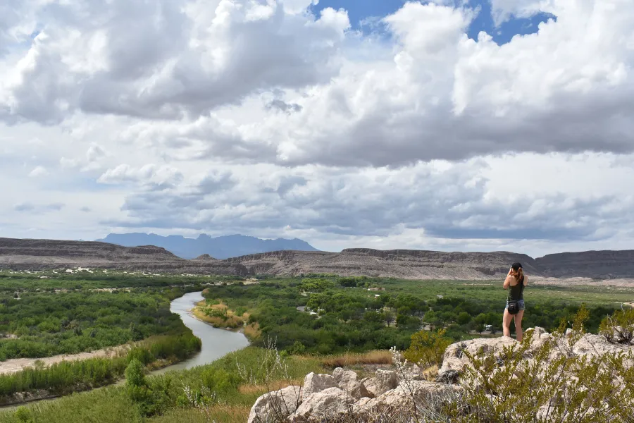 Traveler stands on a rocky overlook above a winding river valley, with wide desert plains and dramatic clouds stretching across the horizon.