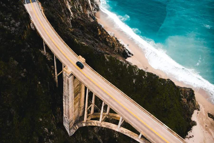 An aerial view of Bixby Bridge spans a rugged canyon along California’s Big Sur coastline. A single dark vehicle crosses the iconic concrete arch bridge, which stretches between steep cliffs above a narrow sandy beach. Below, turquoise ocean waves roll onto the shore, contrasting with the dark rocky bluffs and dramatic coastal landscape.