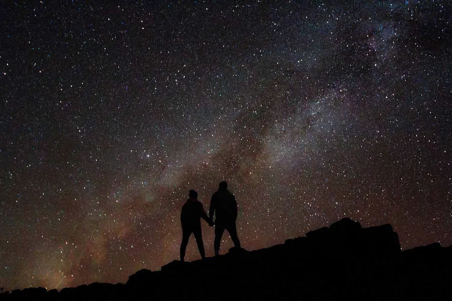 Two people stand silhouetted on a rocky ridge beneath a star-filled night sky and the glowing Milky Way.