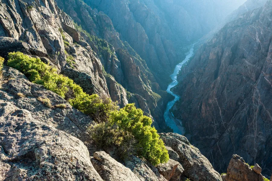 Steep rocky cliffs with patches of green shrubs overlooking the deep, narrow Black Canyon of the Gunnison, with a winding river far below.