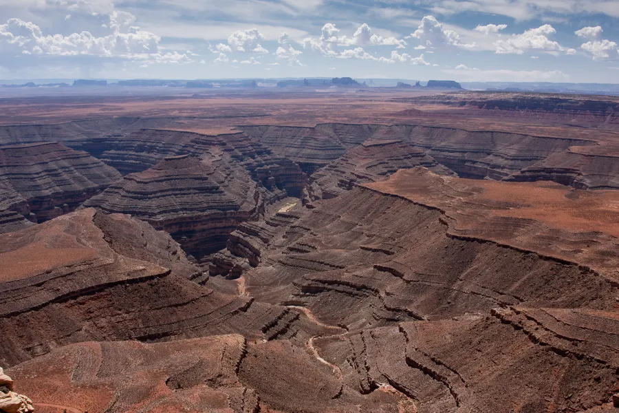 A panoramic view from Muley Point overlooks dramatic layered canyons and mesas stretching across the Utah desert under a sky filled with scattered clouds.