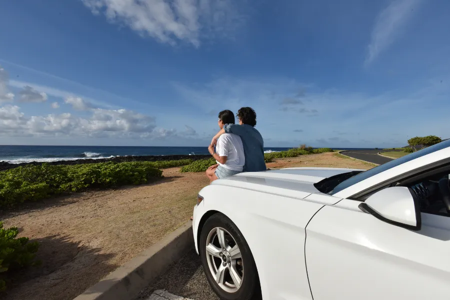 Two travelers sit on the hood of a white car beside the ocean, looking out at the coastal road and blue sky.