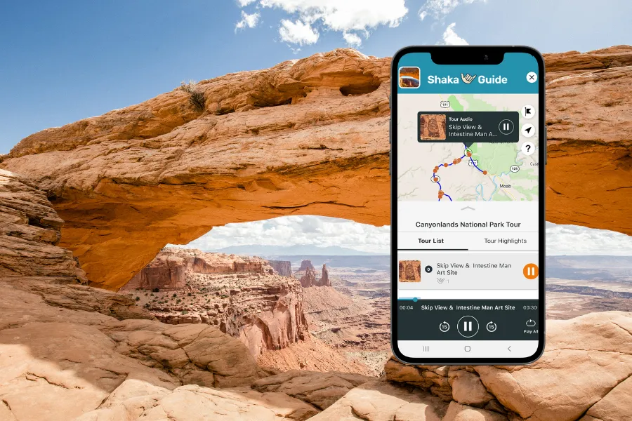 Smartphone displaying the Shaka Guide app beside a wide sandstone arch overlooking a vast red rock canyon landscape.