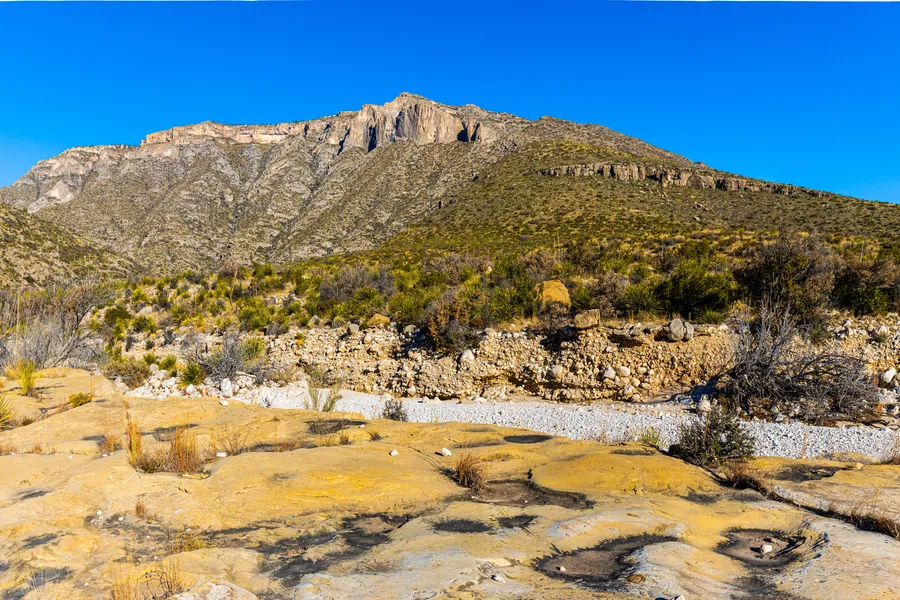 Lichen-covered limestone slabs and ancient stone ruins in the foreground lead the eye toward the rugged, scrub-covered ridgeline of the Guadalupe Mountains under a vivid blue sky along the McKittrick Canyon Trail.