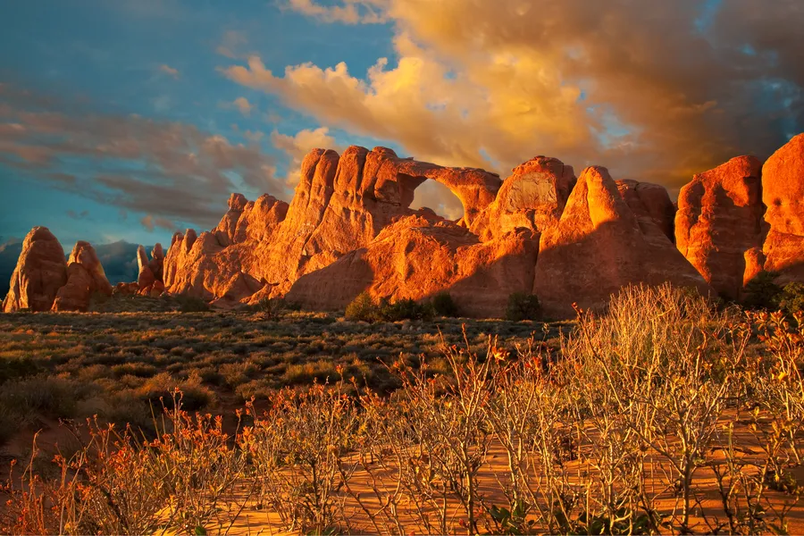 Skyline Arch glows fiery orange in dramatic sunset light at Arches National Park, surrounded by jagged sandstone fins and desert scrubland beneath a stormy, golden-lit sky.