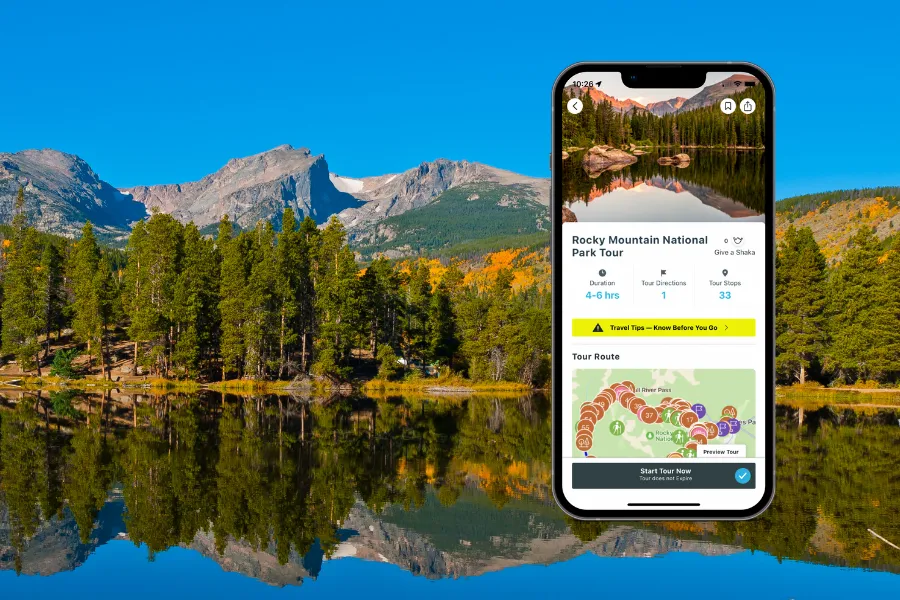 Smartphone displaying a tour app over a reflective mountain lake with pine forests, autumn color, and rugged peaks beneath a clear blue sky.