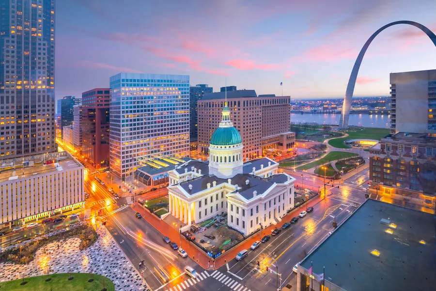 An aerial twilight view of downtown St. Louis showcases the illuminated Old Courthouse with its green dome at center, the Gateway Arch curving against a pink sunset sky, and city streets glowing with light trails along the Mississippi River.