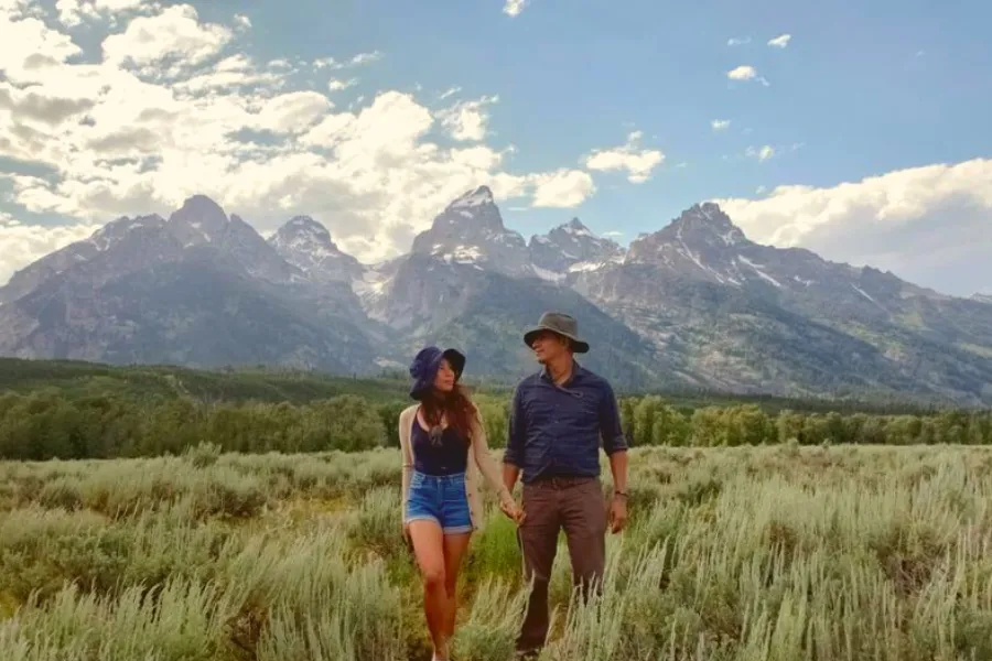 Two travelers walk hand in hand through a grassy field with the jagged Teton mountains rising behind them under a bright sky.
