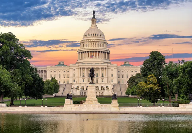 The U.S. Capitol building reflected in the Capitol Reflecting Pool at sunset in Washington, D.C. The iconic white neoclassical dome and grand facade glow warmly against a dramatic orange, pink, and purple sky. In the foreground, the Ulysses S. Grant Memorial — featuring a bronze equestrian statue of General Grant flanked by lion sculptures on a tall white stone pedestal — is centered between the pool and the Capitol steps. Lush green trees frame both sides of the scene.