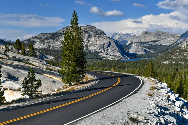 Curving mountain road surrounded by granite rock formations, pine trees, and distant peaks under a clear blue sky.