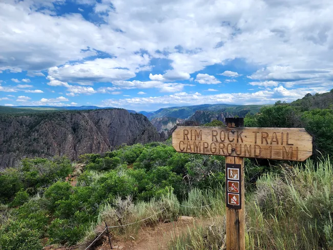 Wooden sign marking Rim Rock Trail and campground direction at Black Canyon of the Gunnison, with dramatic cliffs and lush greenery under a partly cloudy sky.