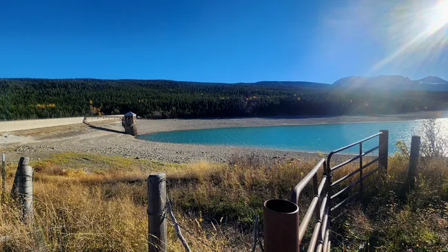 iew of Lake Sherburne Dam with turquoise water, forested hills, and sunlight beaming over mountain peaks.