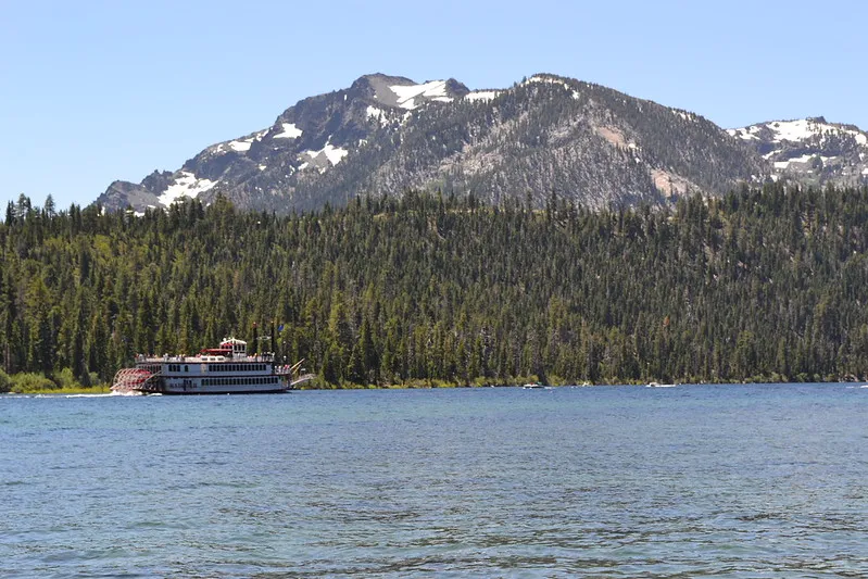 A paddlewheel tour boat moves across Lake Tahoe with dense pine forests and snow-capped mountains in the background.