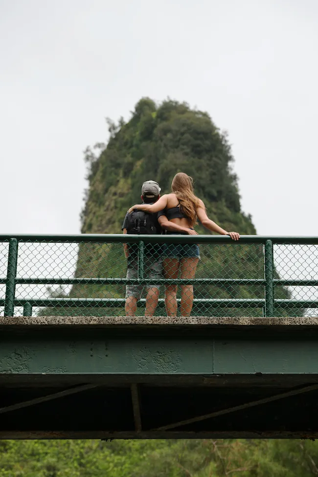 A couple stands on a bridge with arms around each other, looking toward a lush, towering rock formation surrounded by greenery.
