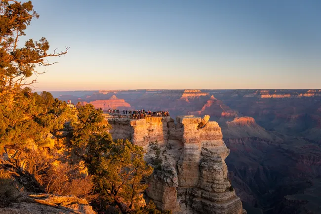 A wide-angle landscape shot of Mather Point at the Grand Canyon’s South Rim during sunset. A large crowd of people is gathered along the railed stone viewing platform, which juts out into the canyon on a layered limestone cliff. The rock face is illuminated with a vibrant golden-orange glow from the setting sun. In the background, the vast canyon stretches toward a soft, hazy horizon under a clear, pale blue sky.