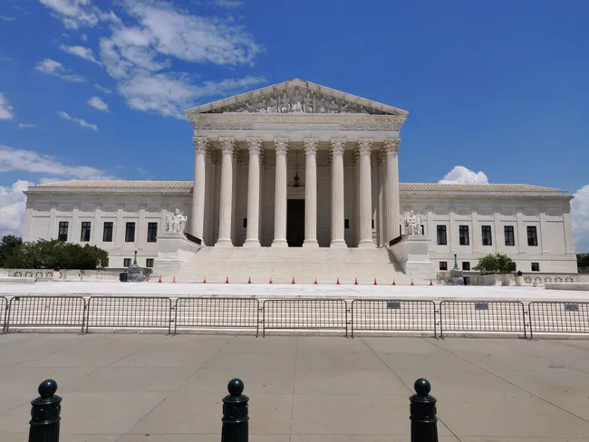 A front-facing view of the United States Supreme Court building in Washington, D.C. The imposing neoclassical white marble structure features a grand pediment with sculptural relief carvings, supported by a row of tall Corinthian columns. Wide stone steps lead up to the entrance, lined with orange traffic cones. Allegorical statues flank the staircase on either side. A temporary metal security fence and bollards line the perimeter at street level. The building is set against a vivid blue sky with scattered white clouds.