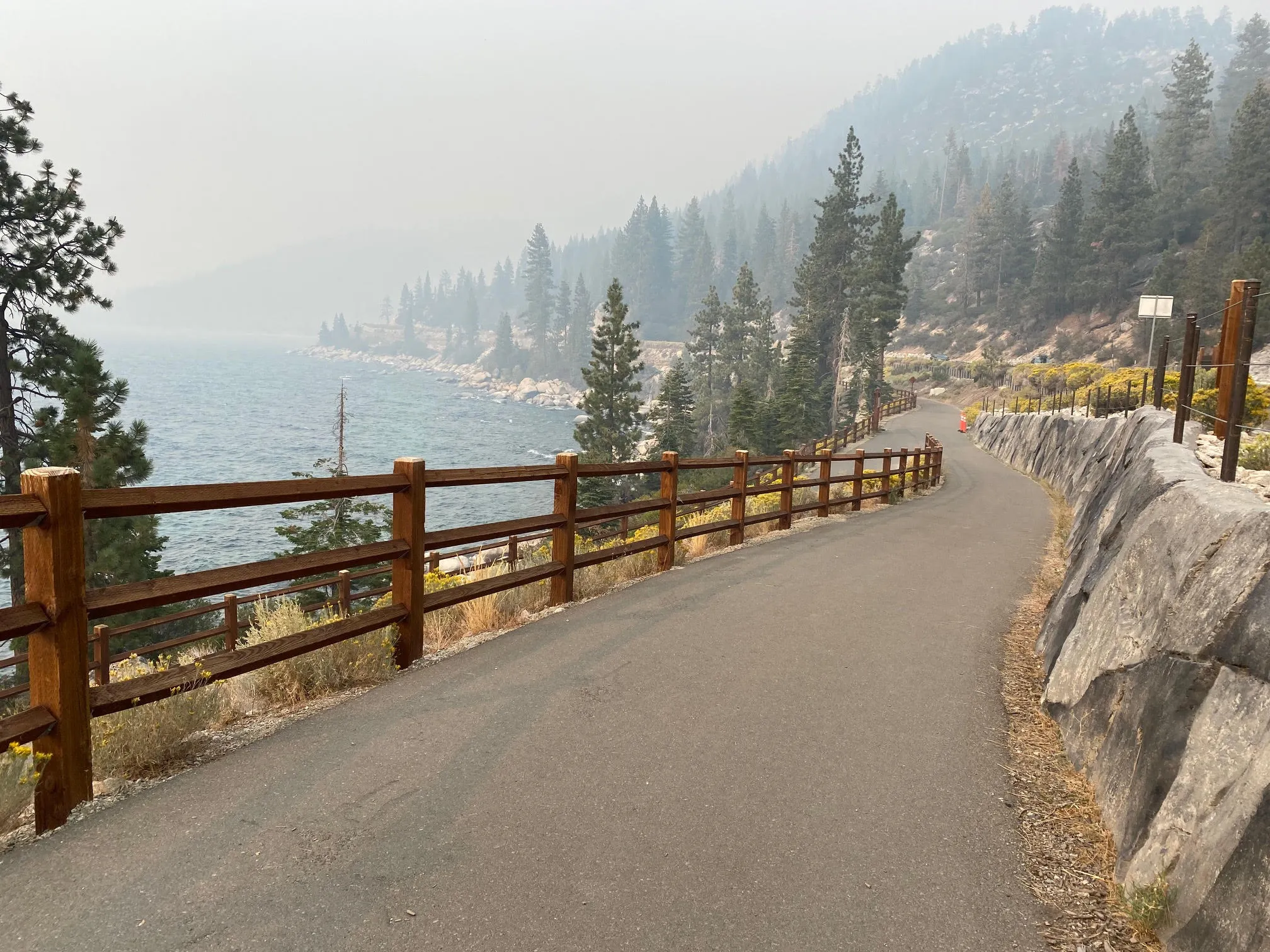 Paved walking trail along Lake Tahoe’s rocky shoreline lined with pine trees and wooden fences under hazy skies.