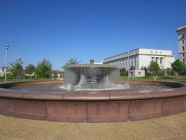 A circular fountain with a wide, mushroom-shaped cascading water feature set in a round red granite basin at Spirit of Justice Park in Washington, D.C. The fountain is centered on a gravel plaza surrounded by a neatly maintained lawn with young trees. A large neoclassical government building with rows of tall windows is visible in the background to the right, under a clear, deep blue sky.