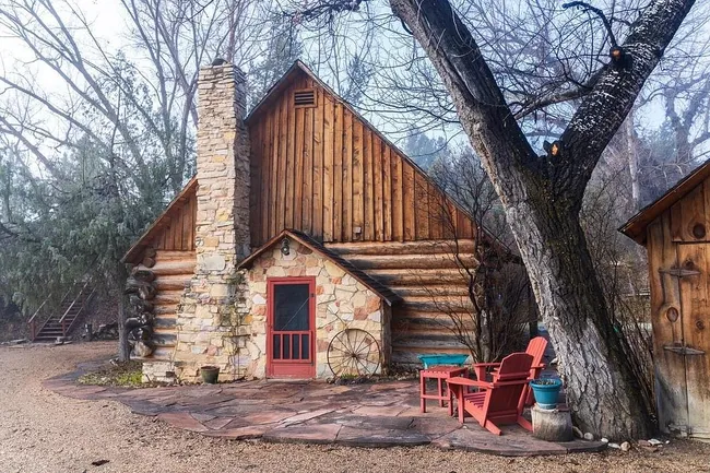 Cozy rustic log cabin with stone chimney, red door, and Adirondack chairs under leafless trees