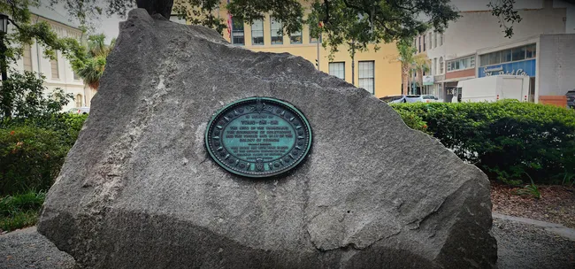 Historic plaque on a large rock in Wright Square, surrounded by greenery and buildings in Savannah, Georgia.